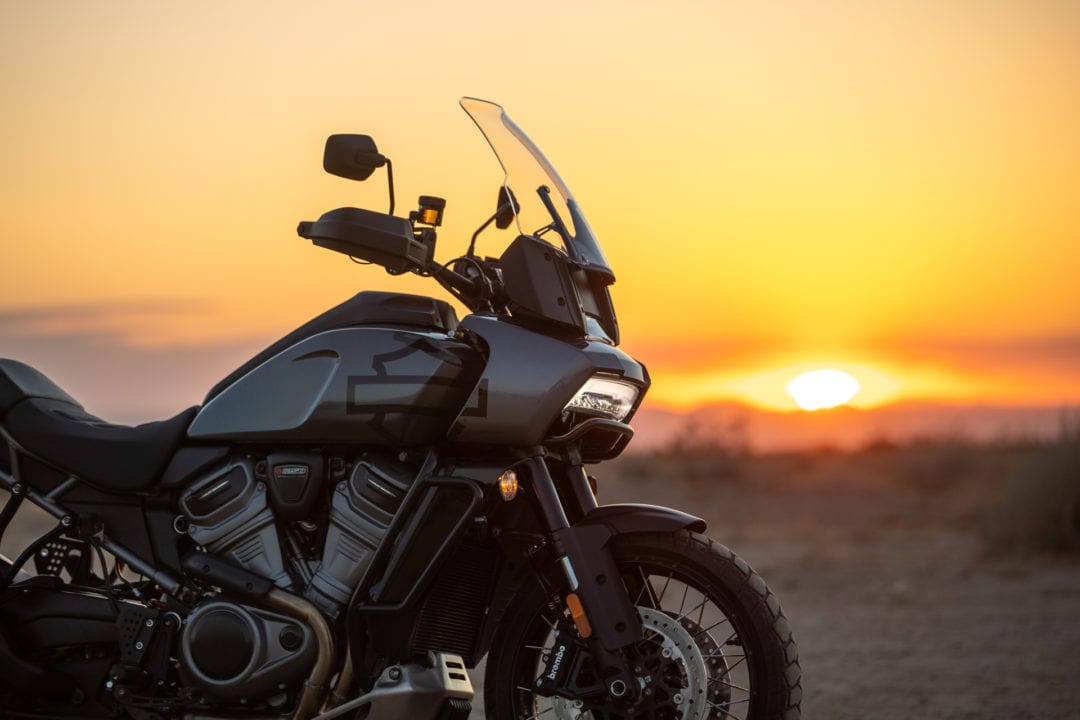 A dark gray Harley-Davidson motorcycle parked in the desert with a sunset behind it. 