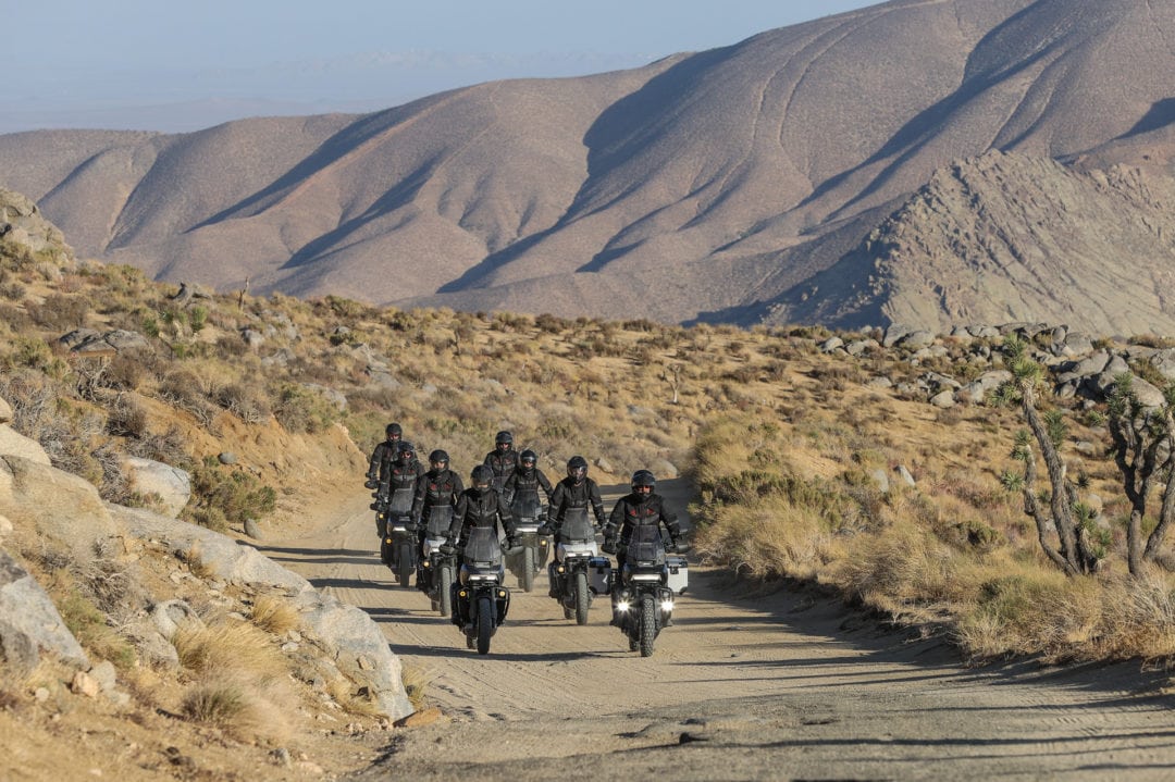 A group of eight riders wearing dark gear riding motorcycles in formation in a desert landscape. 