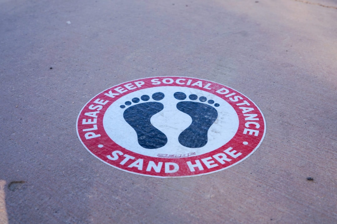 A sign of two feet on the ground reads: "Please keep social distance. Stand here."