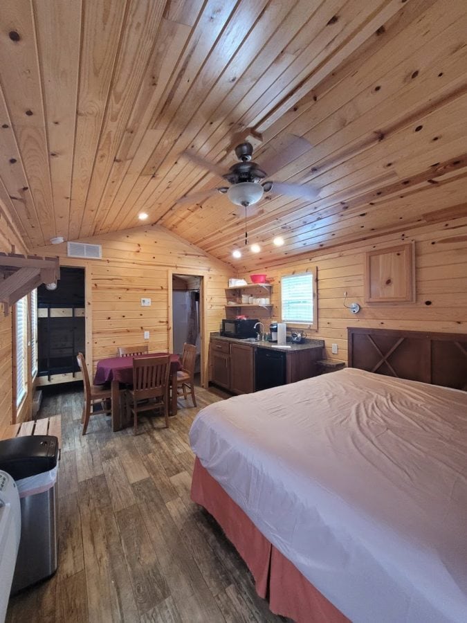 Interior view of inside the cabin with wooden floors and wood paneling on the inside, small kitchenette, table and chairs, and bed with a white sheet