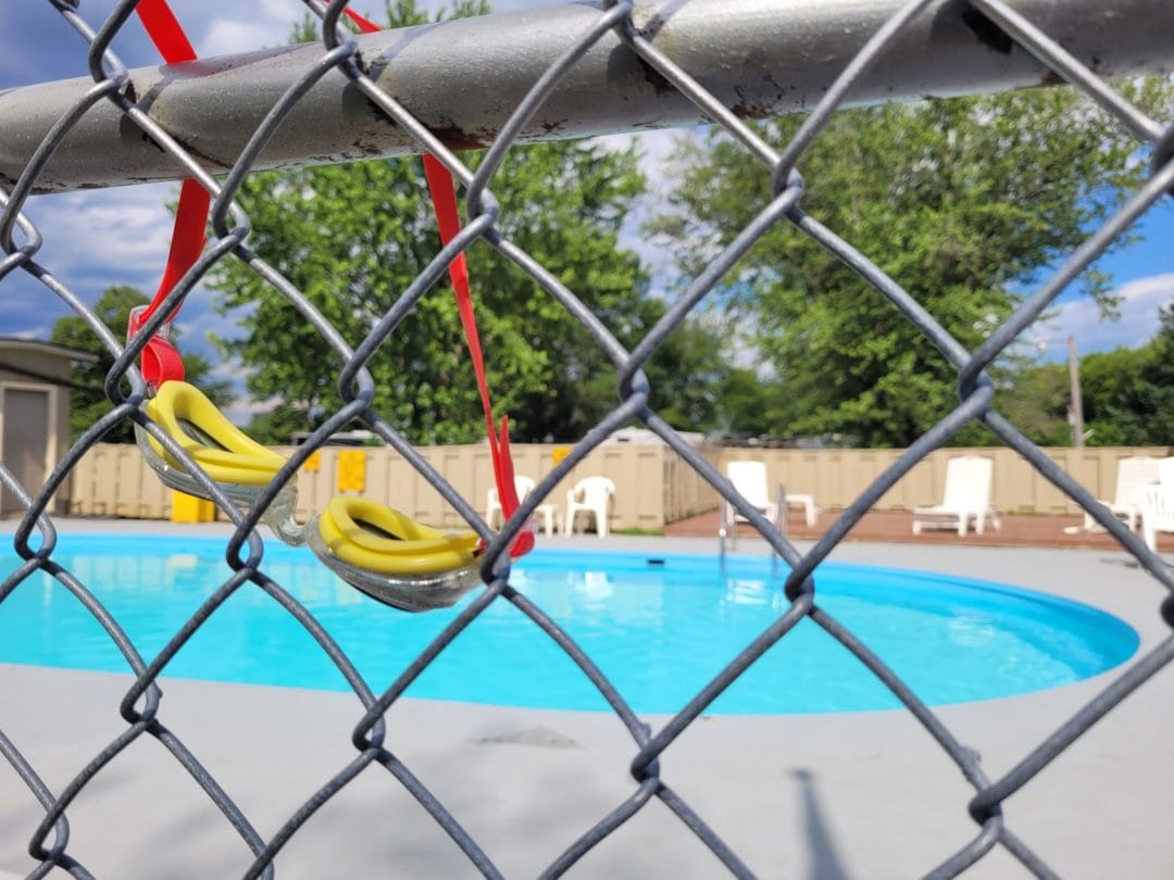 View of a clear blue swimming pool through a chain link fence with yellow swimming googles hanging from the fence