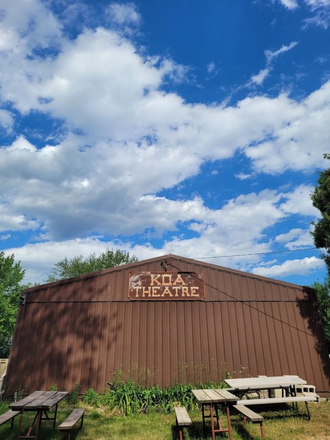 Large, brown barn-like structure with KOA Theatre sign on the side and picnic tables around it