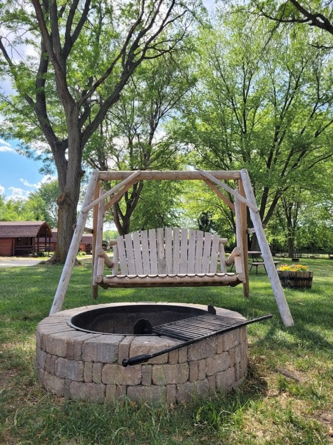 Wooden swing in front of a stone fire pit surrounded by green trees and blue skies
