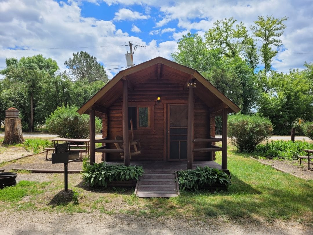 Small wooden cabin on grass surrounded by bushes and trees with porch and swing