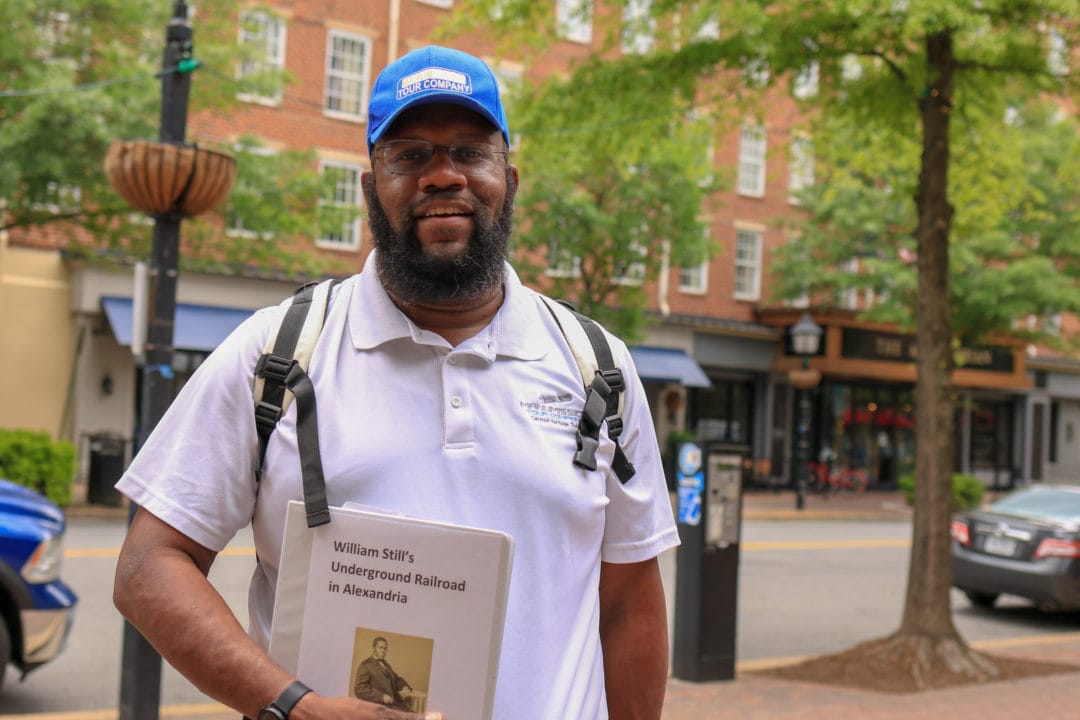 a black man wears a white polo and blue hat and holds a binder that says "william still's underground railroad in alexandria"