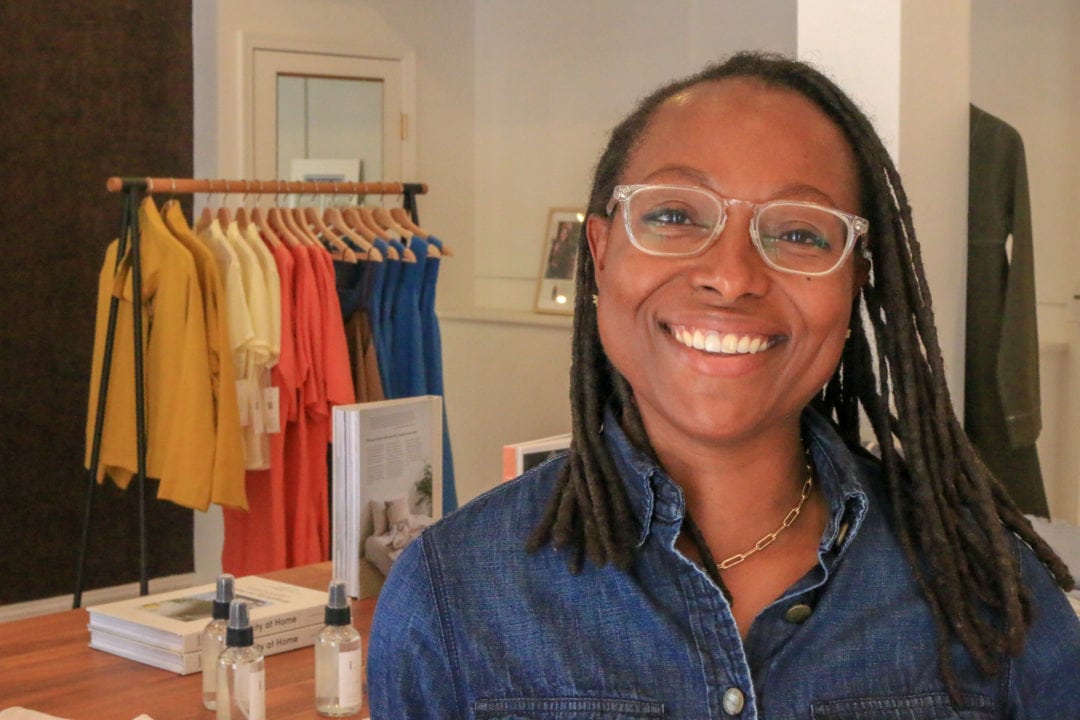 a black woman wears clear glasses and a blue shirt and smiles in front of a rack of clothes