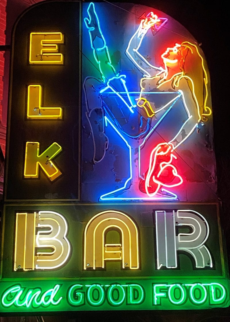 a neon sign that says "Elk Bar and Good Food" with a woman sitting in a cocktail glass