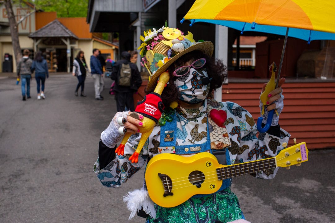 a woman wears a facemask, sunglasses and a hat and carries two rubber chickens and a ukulele