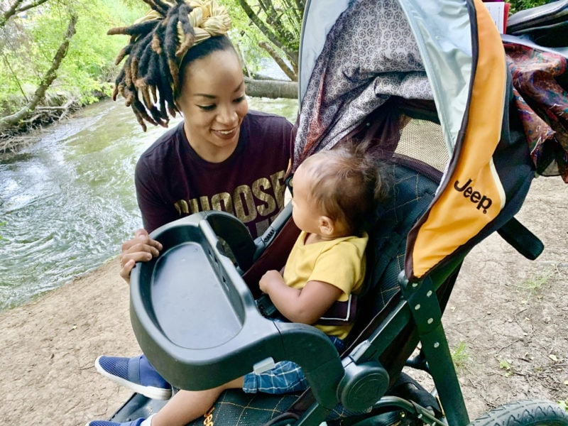 a Black woman kneels near a stroller and smiles at her baby