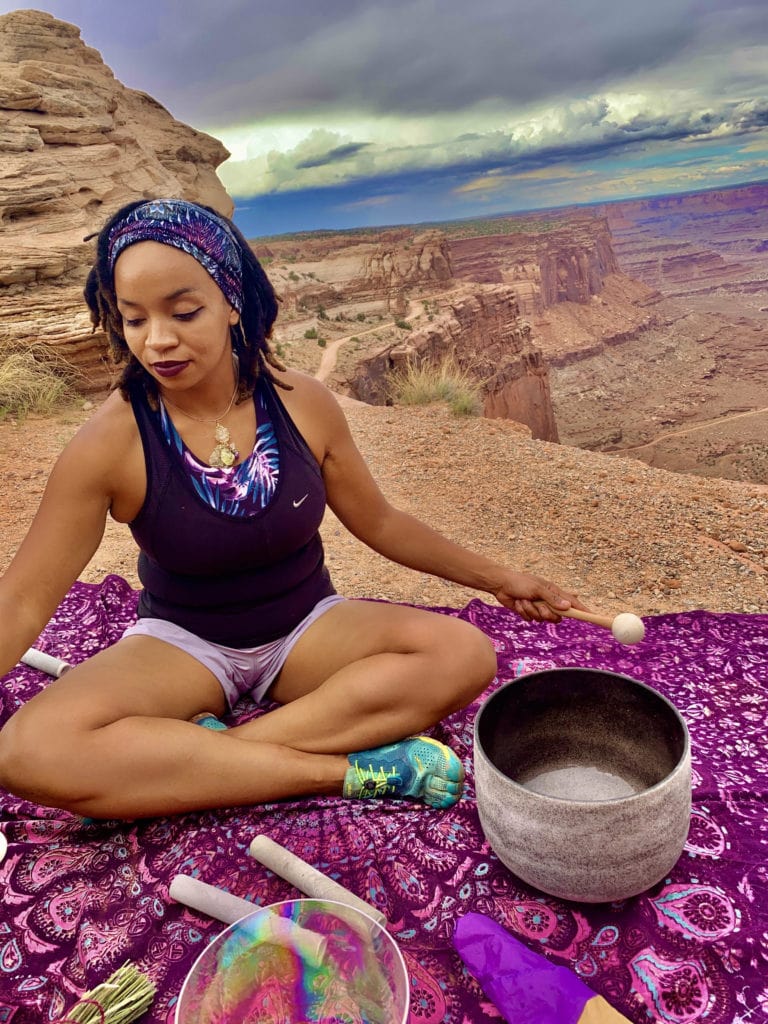 a black woman sits cross-legged on blanket near a scenic desert overlook performing a sound healing