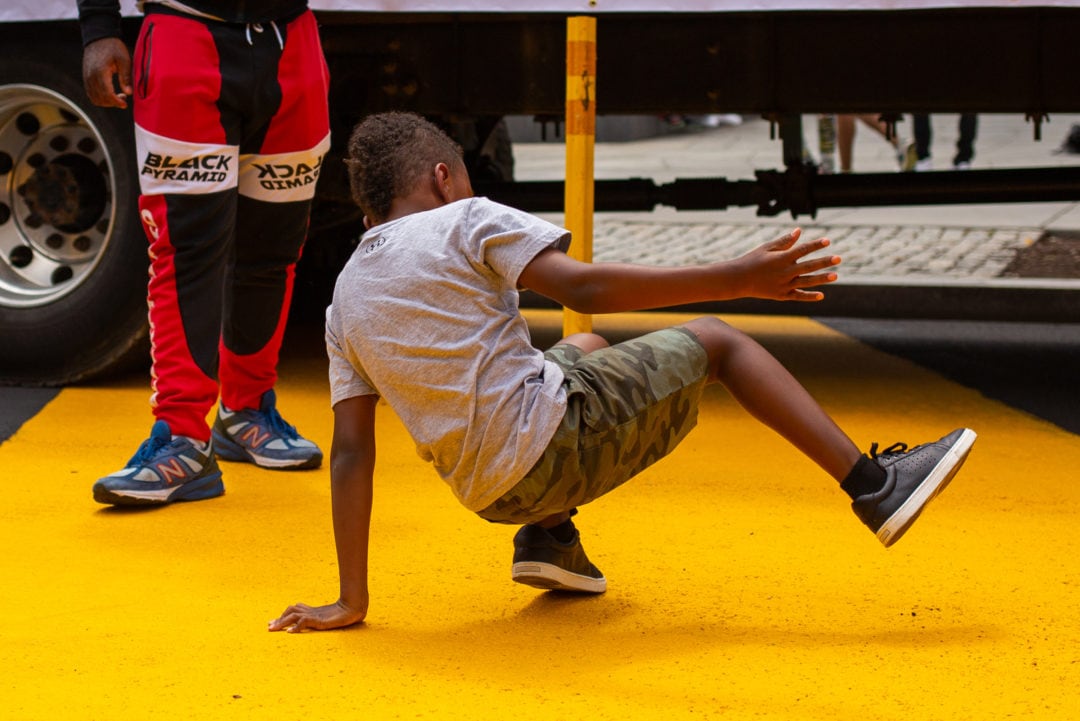 a Black boy shows off his dance moves on a painted yellow street