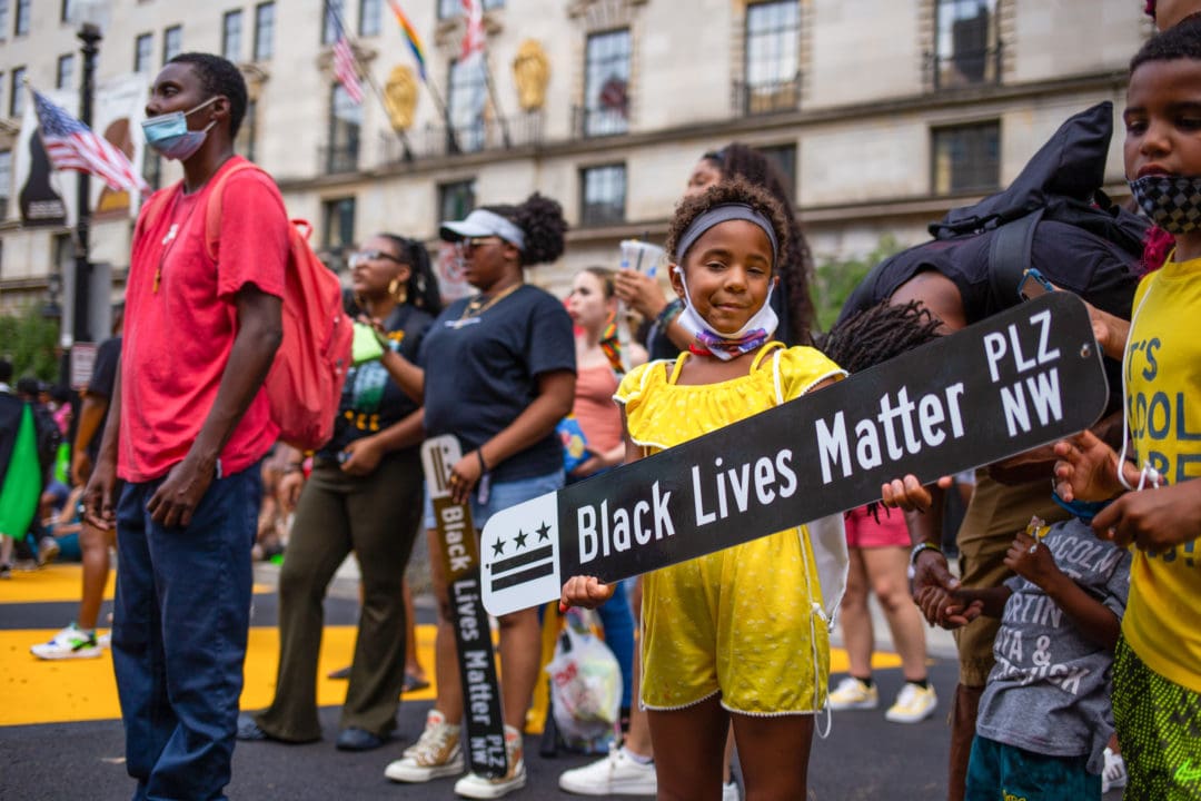 a young Black girl holds a street sign that says "Black Lives Matter Plaza NW"
