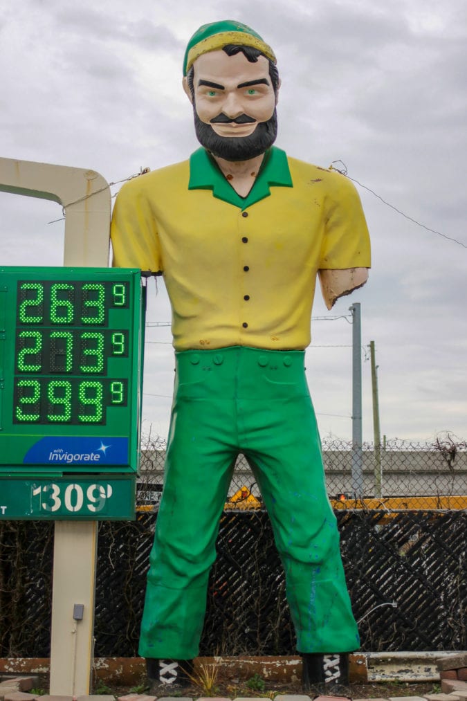 an armless muffler man stands at a gas station wearing a yellow shirt and green pants