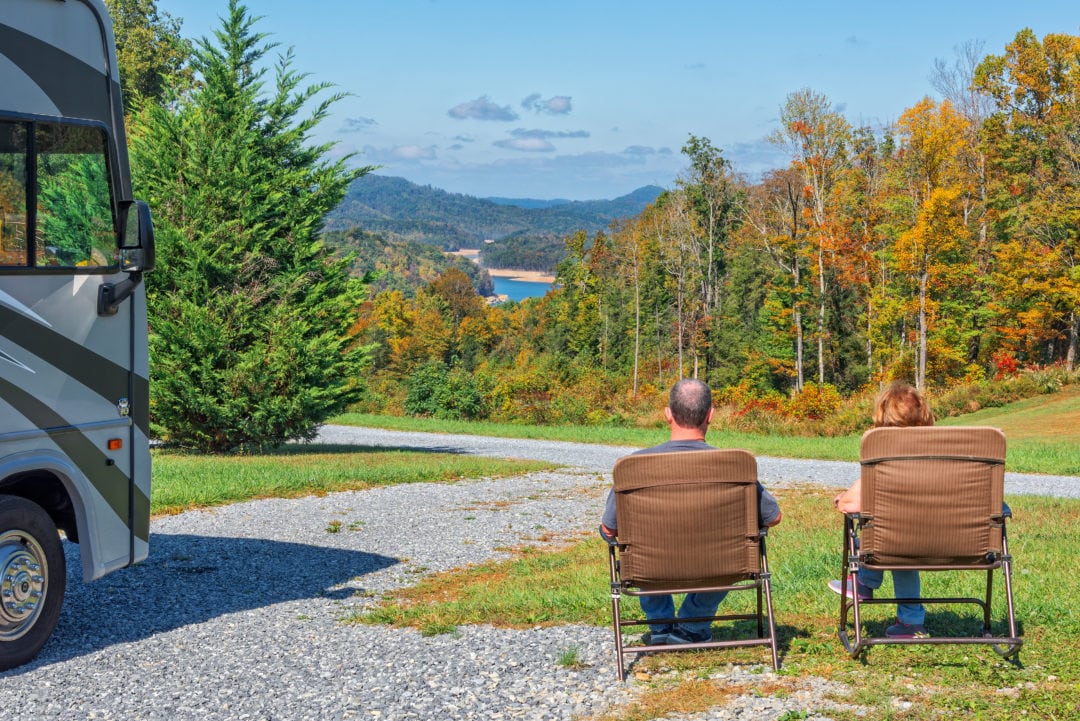 Two white people sit in folding chairs looking at a forest next to an rv