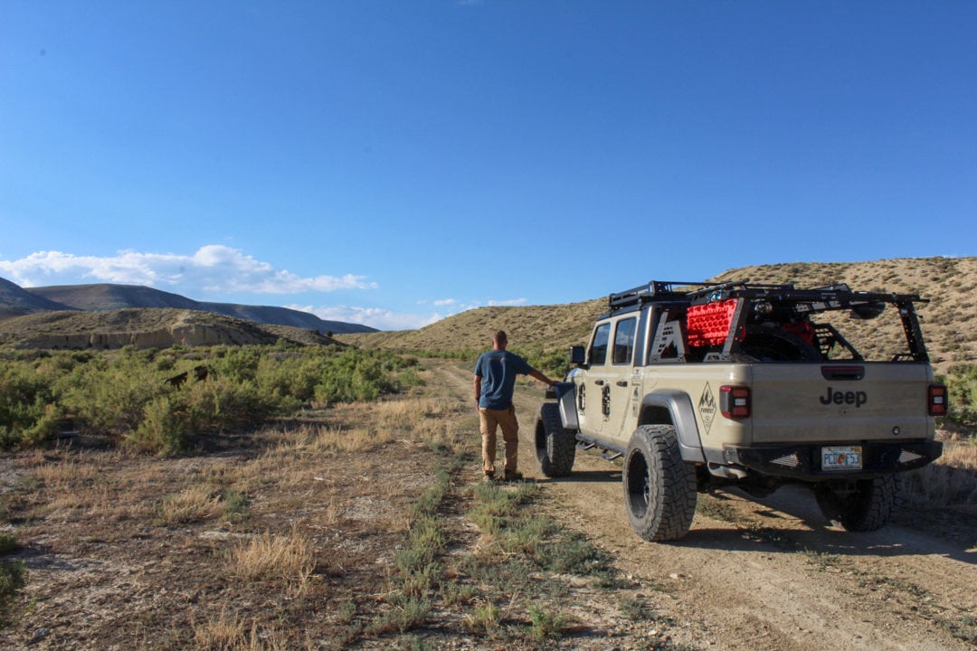A man with his back against the camera stands near a jeep on an empty trail