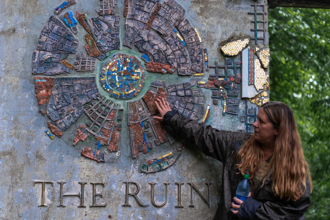 a white woman with long wavy hair touches a circular mosaic sign that says "the ruins"