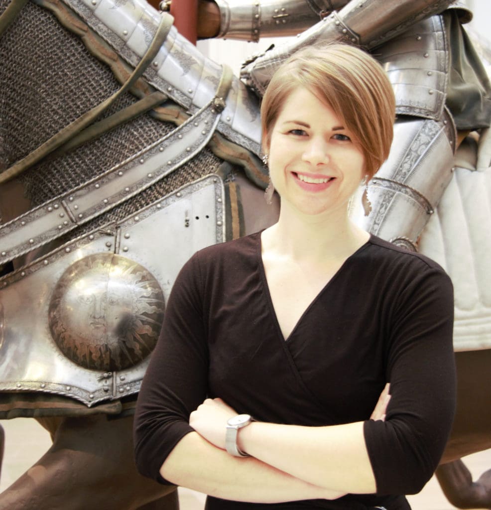 A white woman with short brown hair stands with her arms crossed smiling in front of a suit of armor