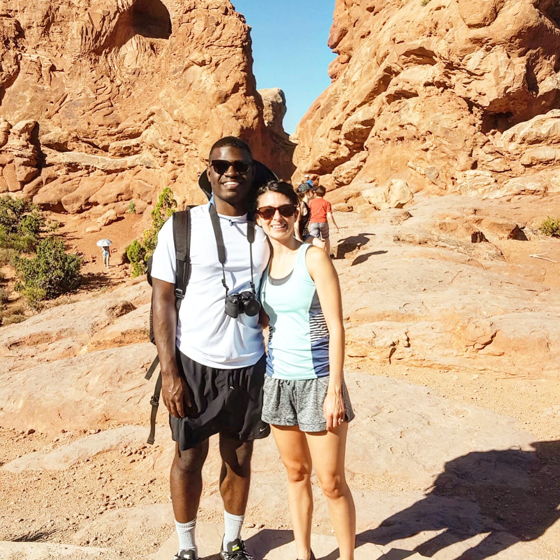 a black man with sunglasses and a camera around his neck poses with a white woman wearing sunglasses on a hiking trail in the desert