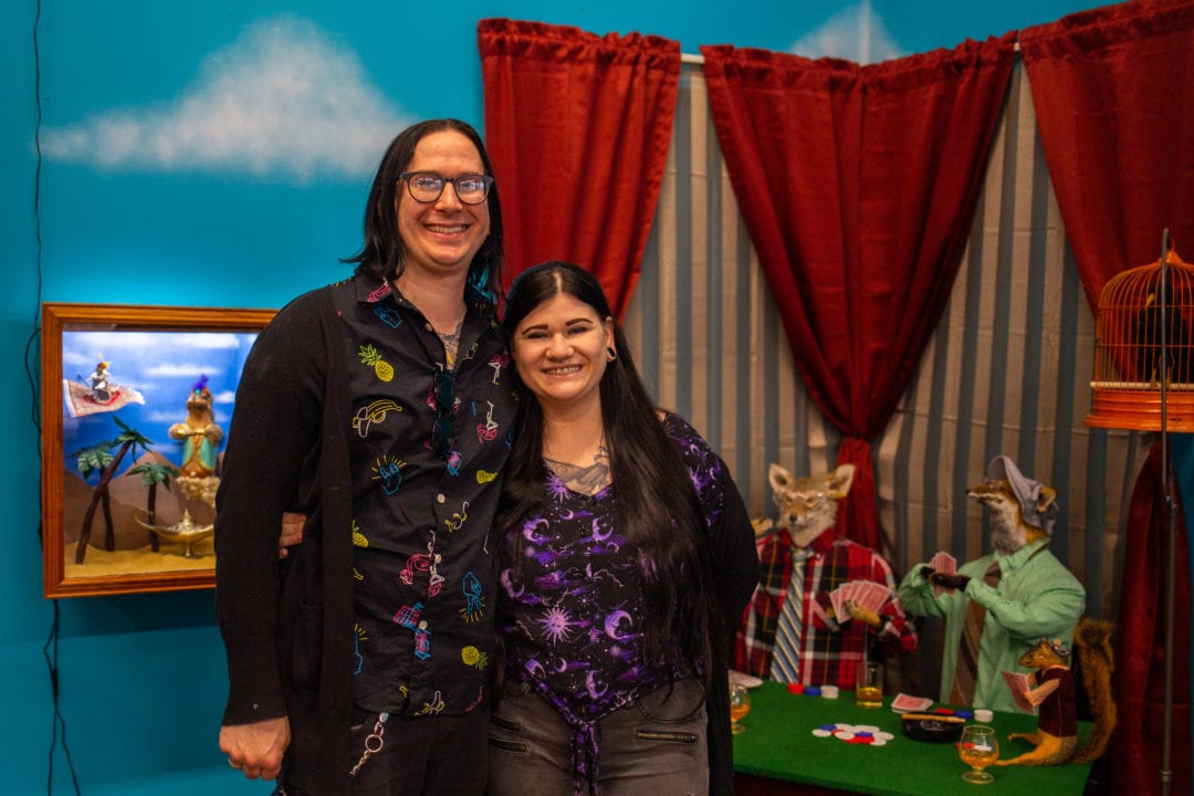 A white man and woman stand smiling in front of a blue wall and taxidermy dioramas
