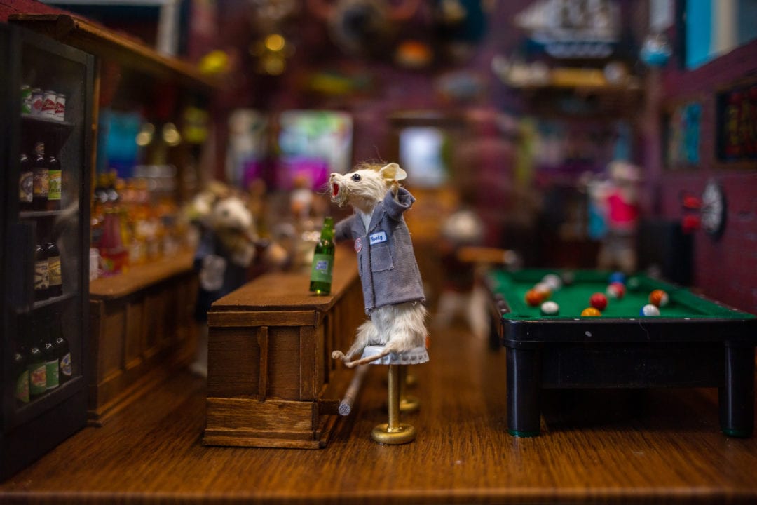 A taxidermy mouse sits at a bar stool and drinks a tiny beer next to a pool table