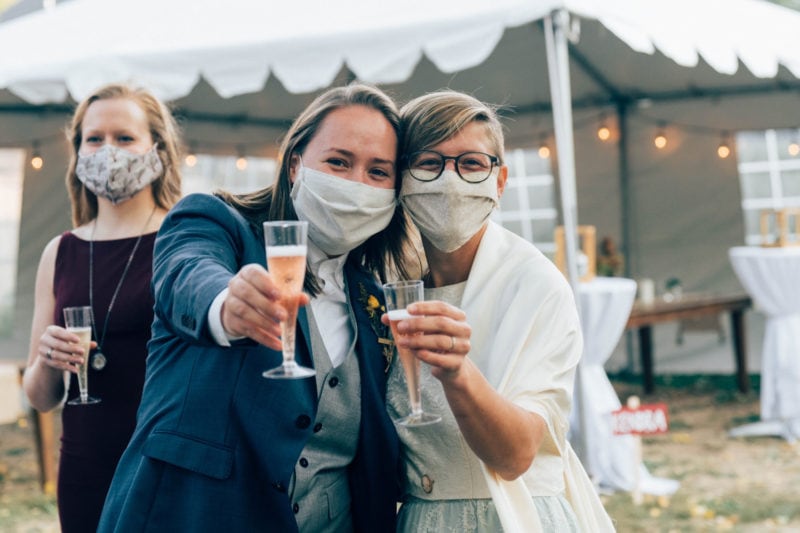 Two brides wearing masks and smiling hold up champagne flutes to the camera