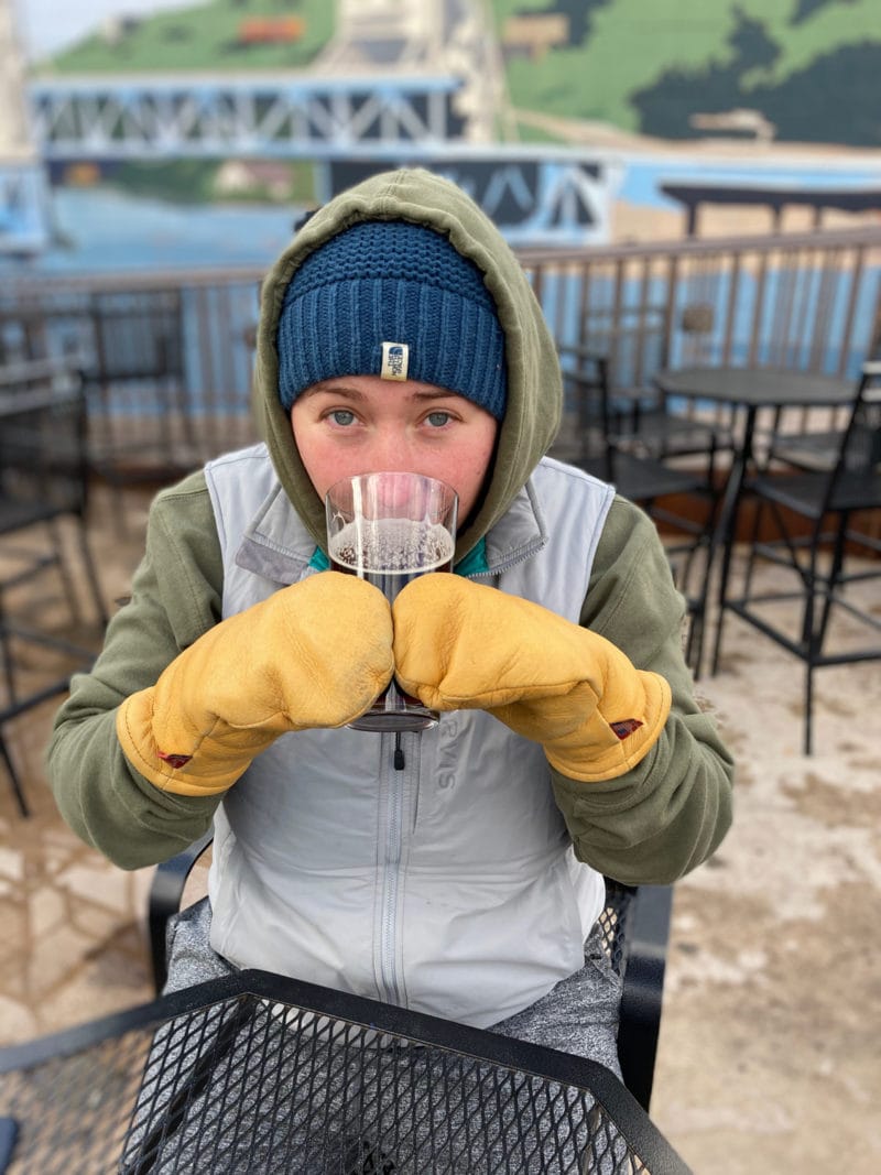 A person dressed in warm clothes and gloves is drinking a beer held with both hands