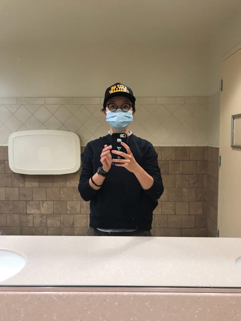 A person wearing a mask taking a selfie in a gas station bathroom mirror