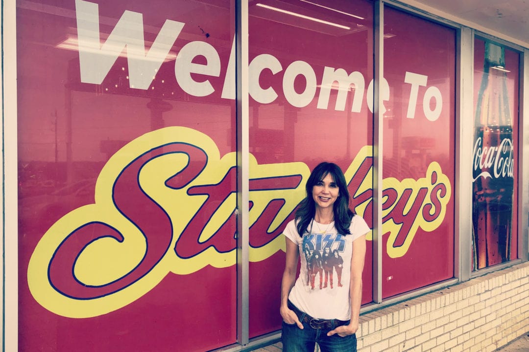 A white woman with brown hair wears a white Kiss tshirt and jeans and stands in front of a sign that says "welcome to Stuckey's"