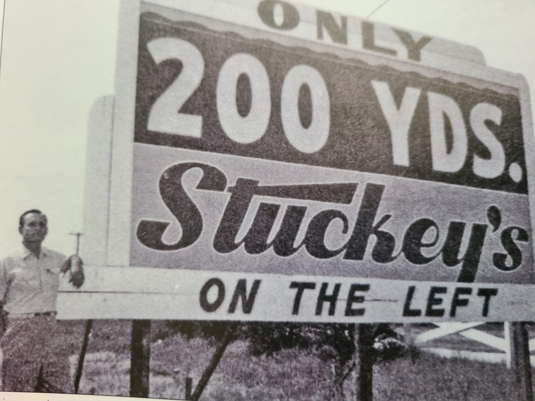 A black and white photo of a man standing next to a billboard that says "only 200 yds. stuckey's on the left"