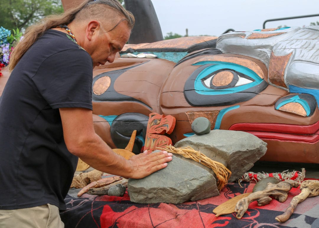 A man with a shaved head places his hand on a rock left near a totem pole