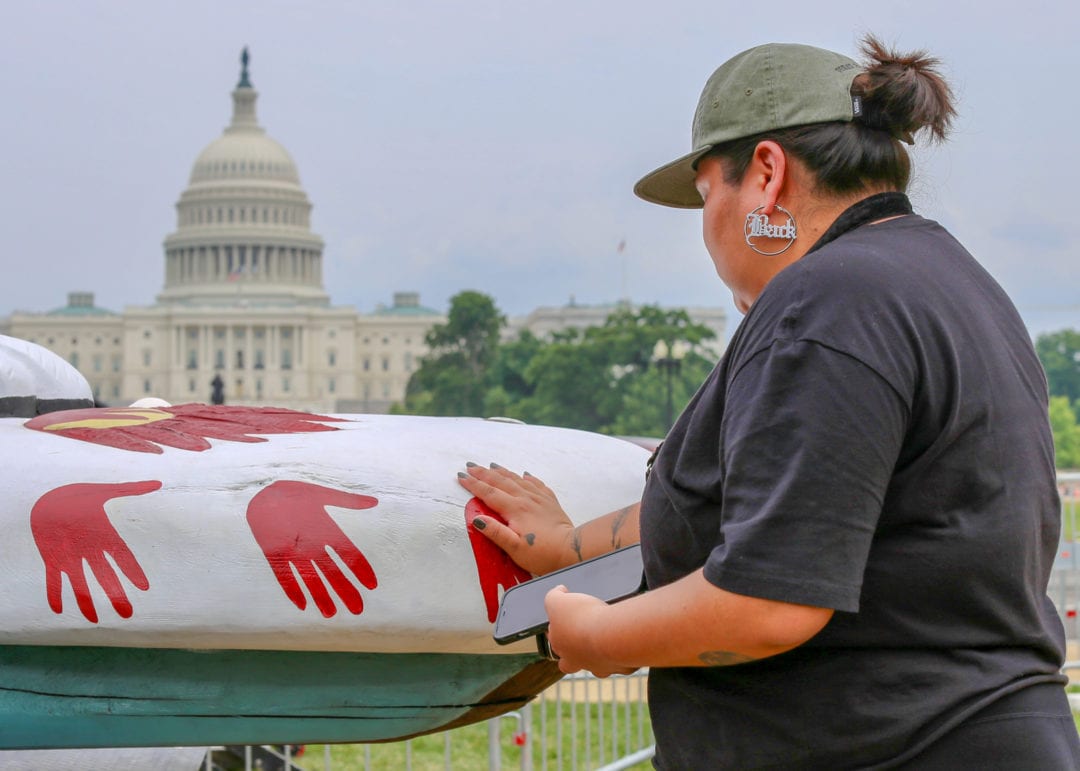 A woman places her hand on a red painted hand print on the totem pole in front of the capitol dome