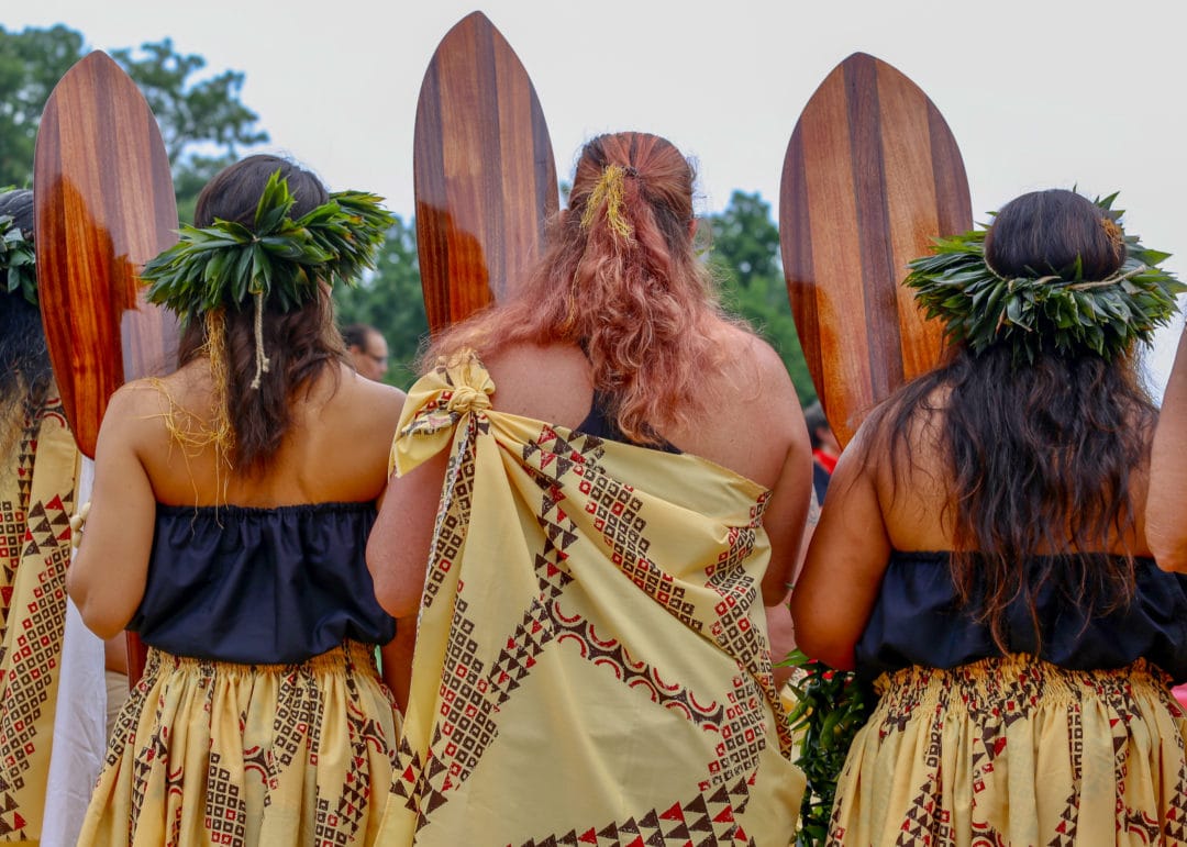 Three women wear draped fabric, head dresses made of green leaves and hold wooden surf boards