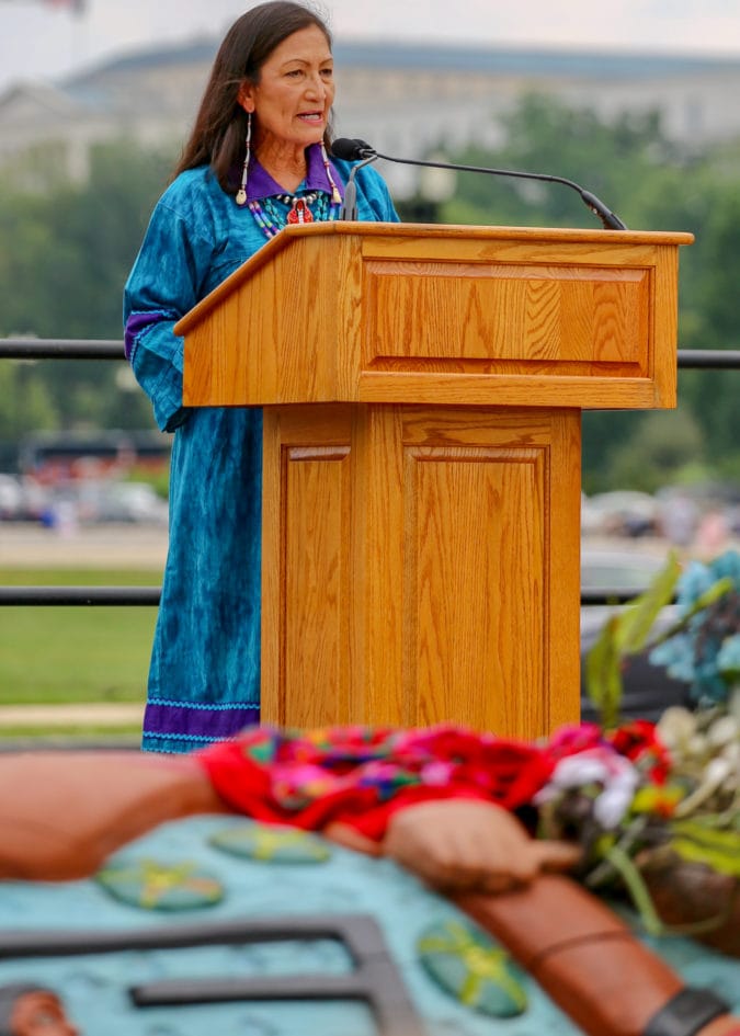 A native woman wearing a bright blue and purple dress and long earrings speaks behind a podium