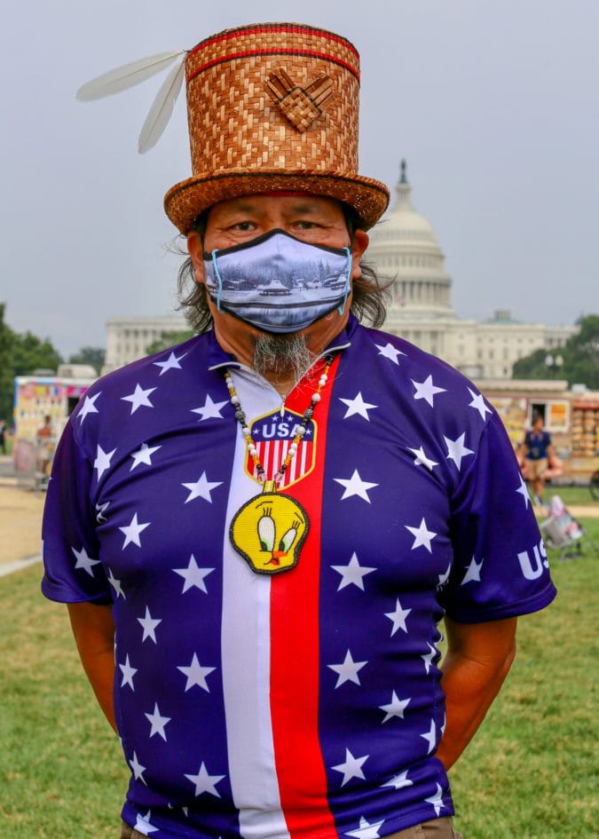 a man in an american flag shirt and straw top hat wears a beaded tweet bird medallion and poses in front of the capitol