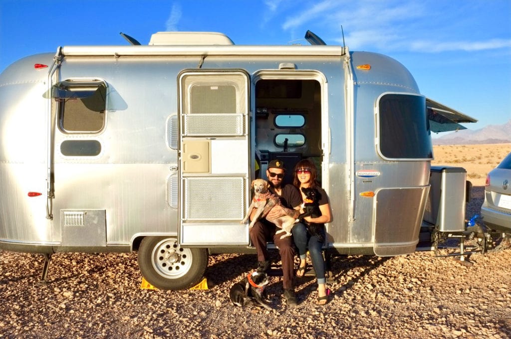A man and a woman seated in the doorway to an Airstream trailer, each holding a dog