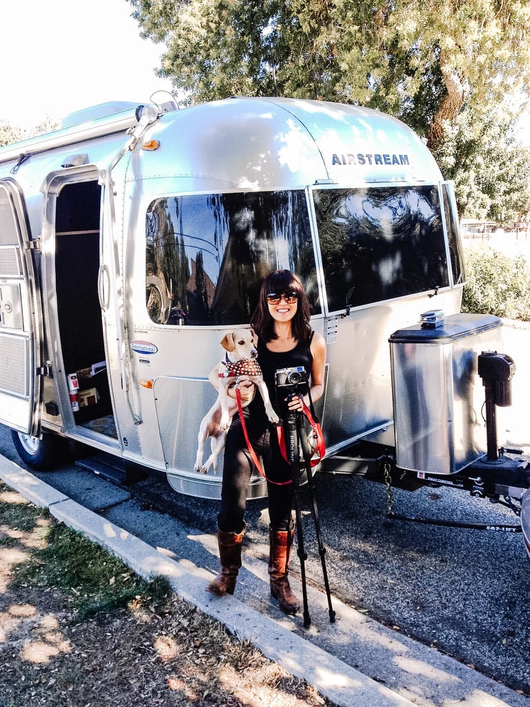 A woman holding a dog and a camera on a tripod, standing in front of an Airstream trailer