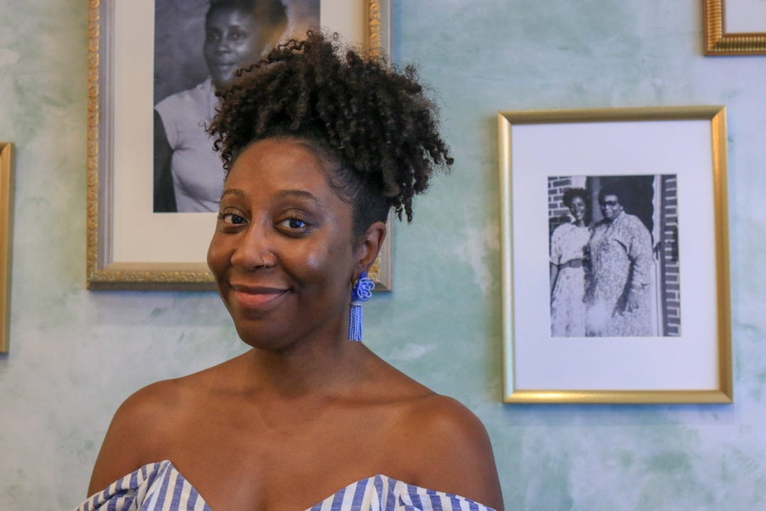 a black woman smiles standing in front of framed photos of her family