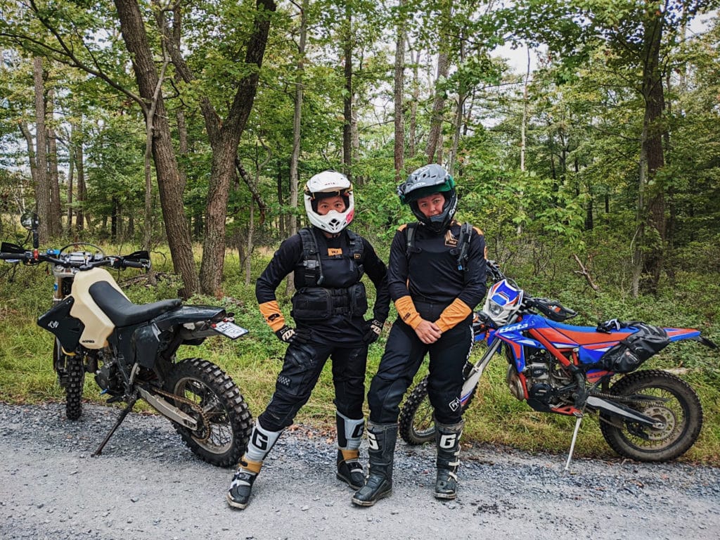 two people in motorcycle gear stand by the side of a road in front of two bikes