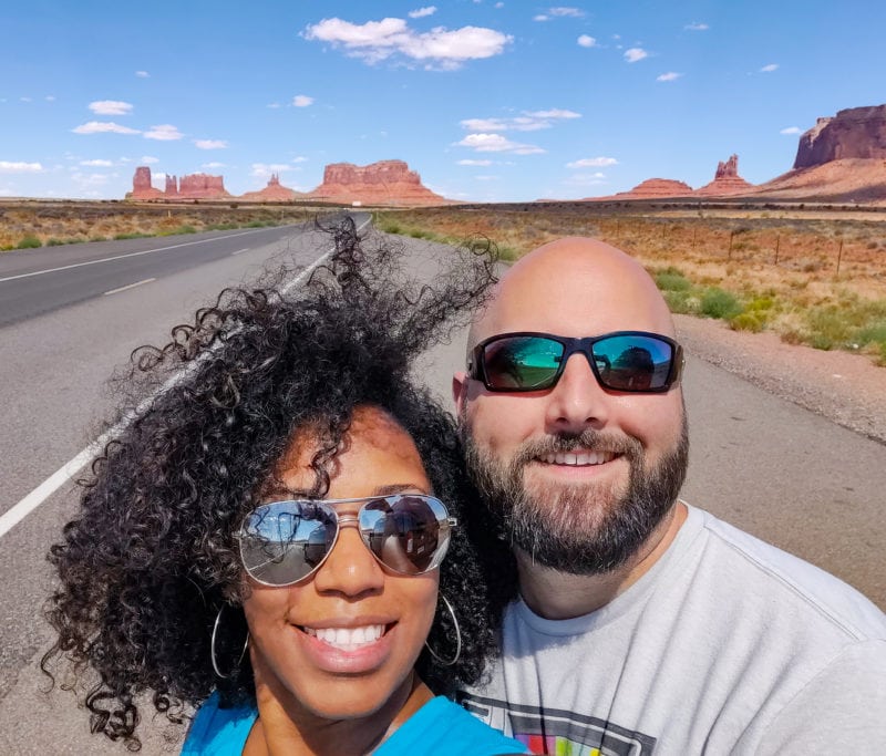 two people pose for a selfie in front of a stretch of road with rock formations and a blue sky