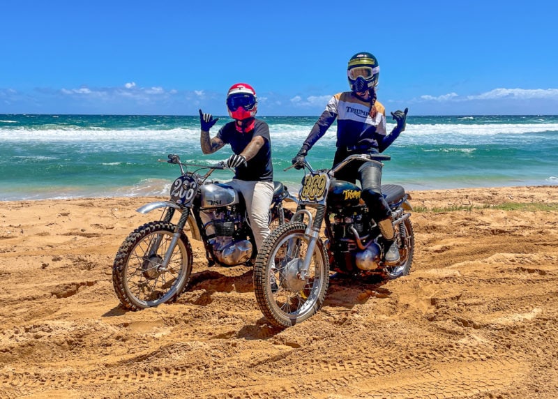 Two people on vintage motorcycles on a sandy beach with a blue sky and ocean in the background