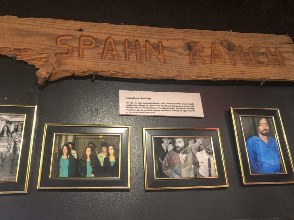 a wooden sign that says "spahn ranch" hangs above photos of charles manson and his "family"