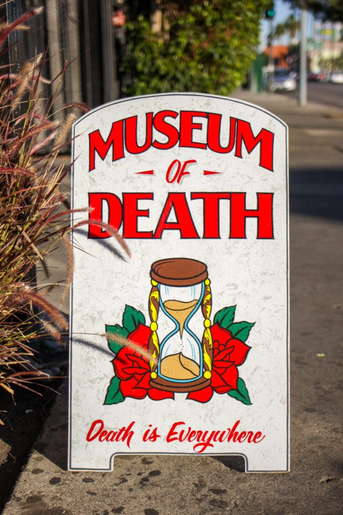 a board sign for the museum of death featuring an hourglass and the words "death is everywhere"