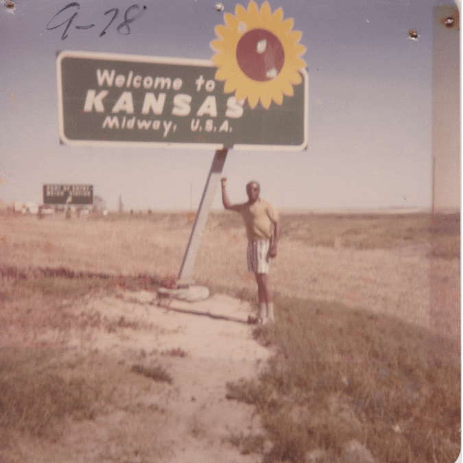 a man poses in front of a welcome to kansas sign