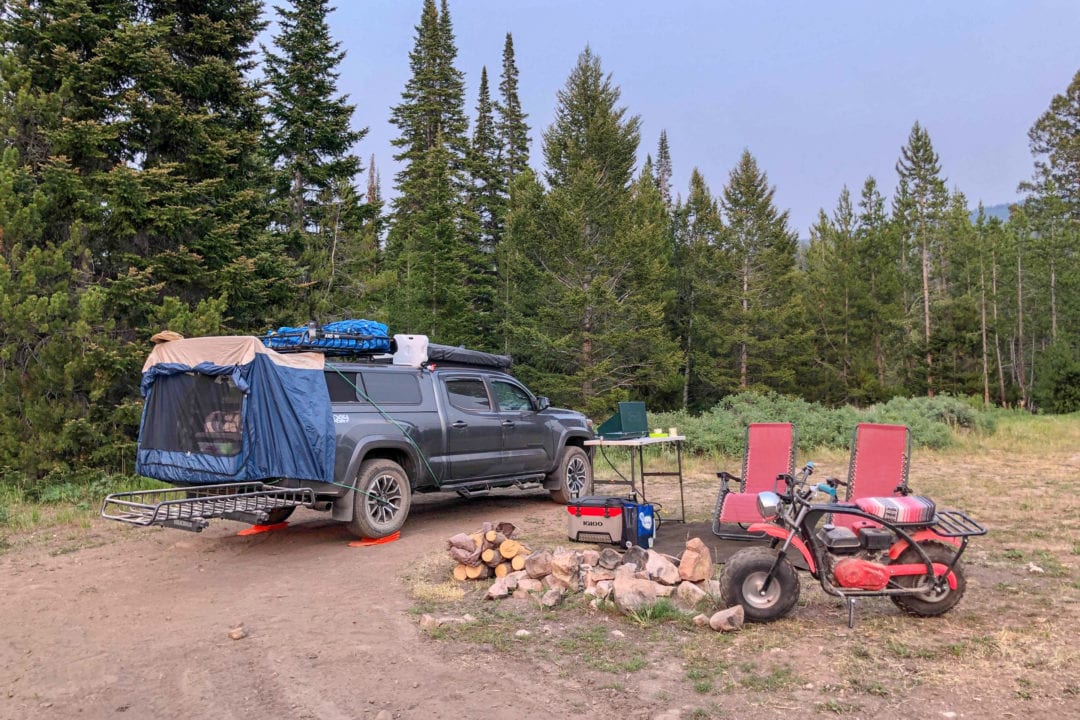 a truck outfitted to camp at a campsite with a firepit, motorbike and two red chairs