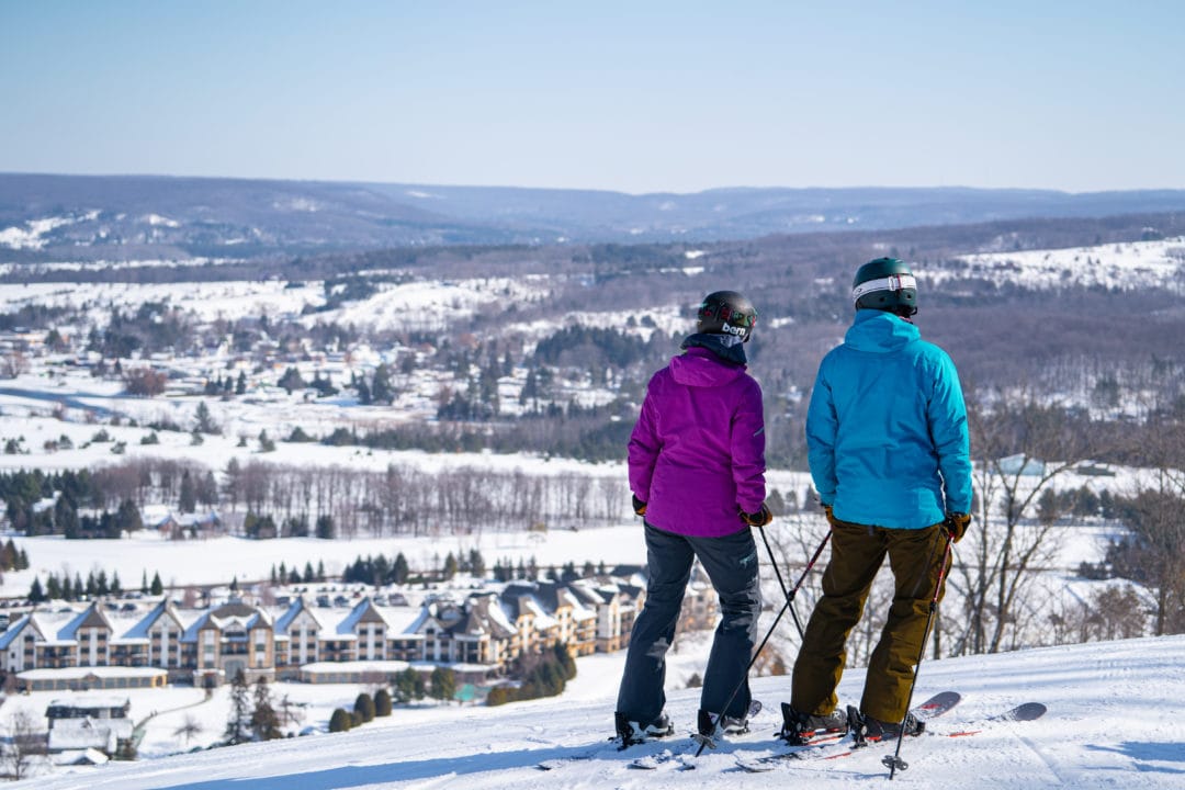 Man and woman standing at the top of a snowy ski hill with their skis and ski poles.
