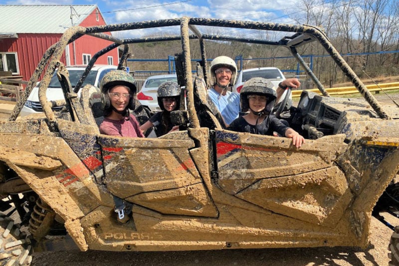 four people wearing helmets and goggles sit in an off-road vehicle covered in mud