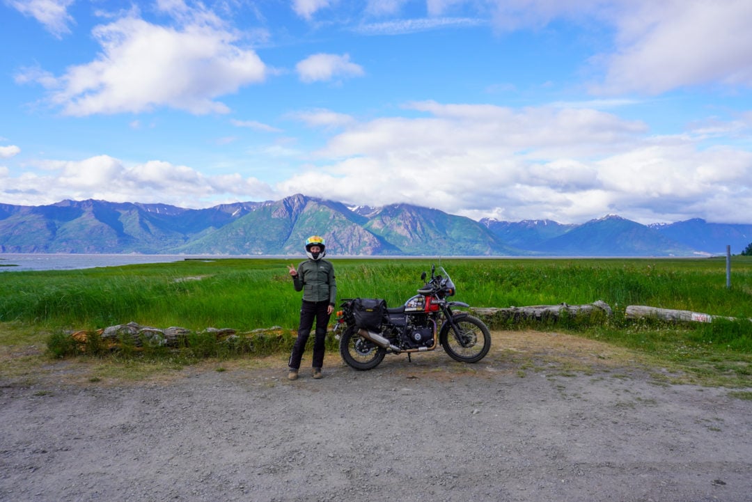 A woman standing next to a motorcycle with mountains in the background
