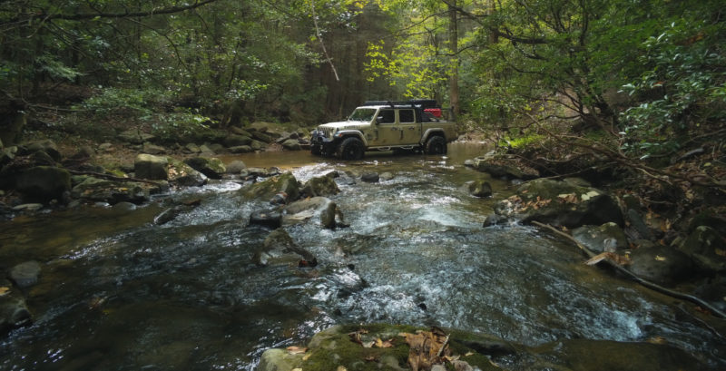 a beige jeep sits in a creekbed surrounded by trees
