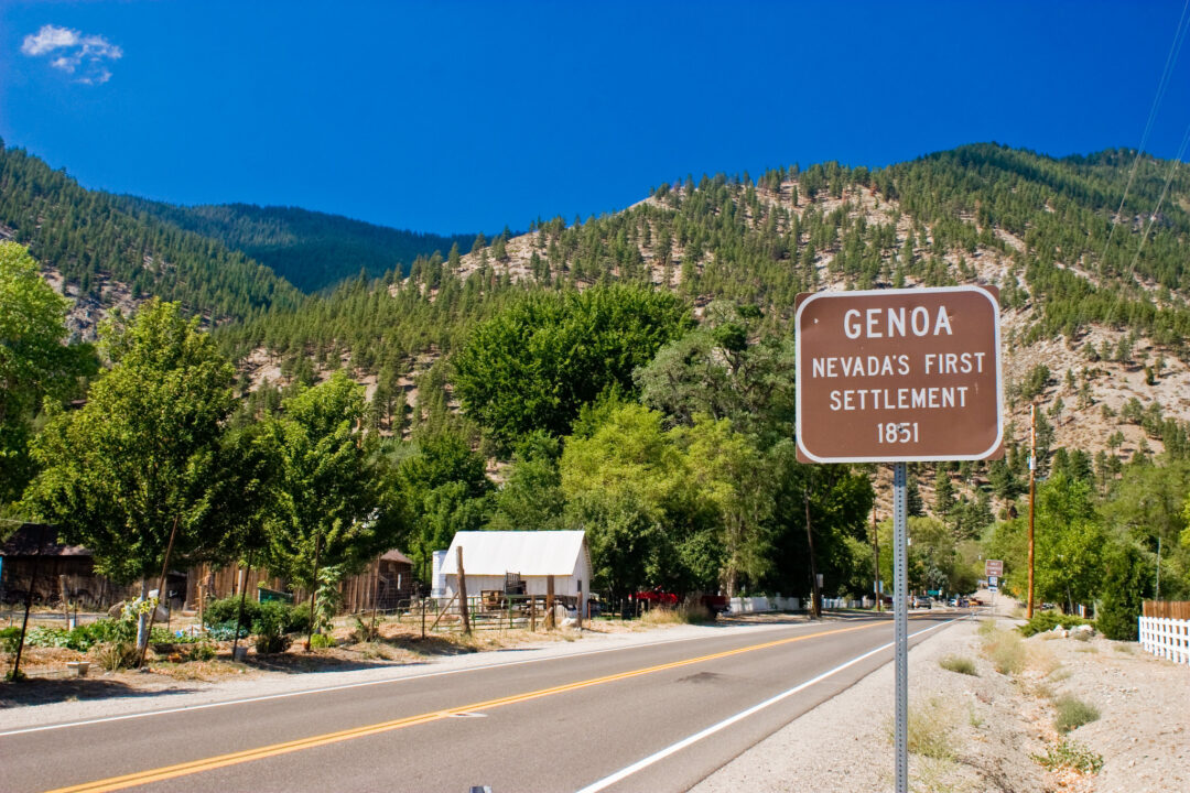 a brown road sign says "genoa nevada's first settlement 1851" next to a road in front of a mountain of green pine trees and a blue sky
