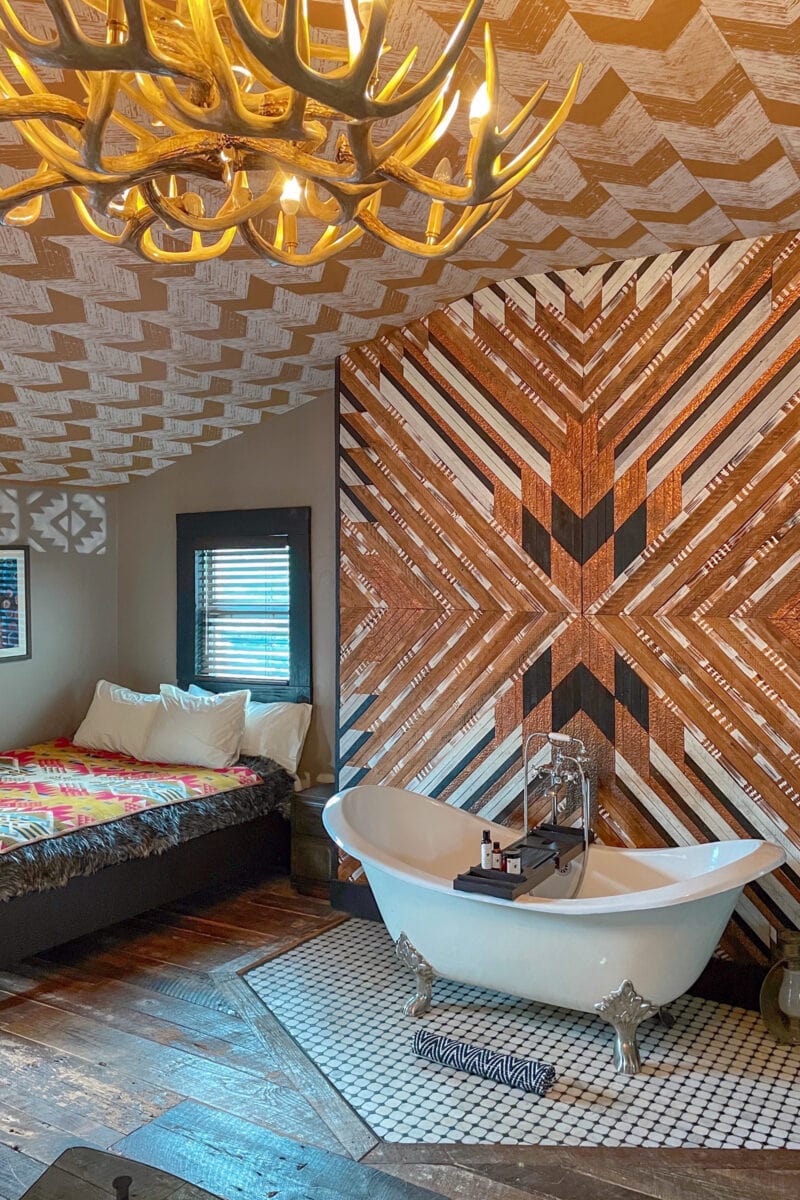 a hotel room with an antler chandelier patterned walls, and a bed and claw foot tub
